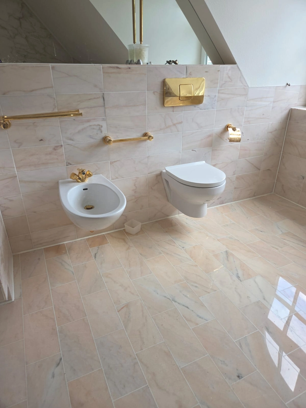 Wandhängendes WC und Bidet mit goldfarbenen Armaturen und heller Marmorfliesenoptik – Badrenovierung