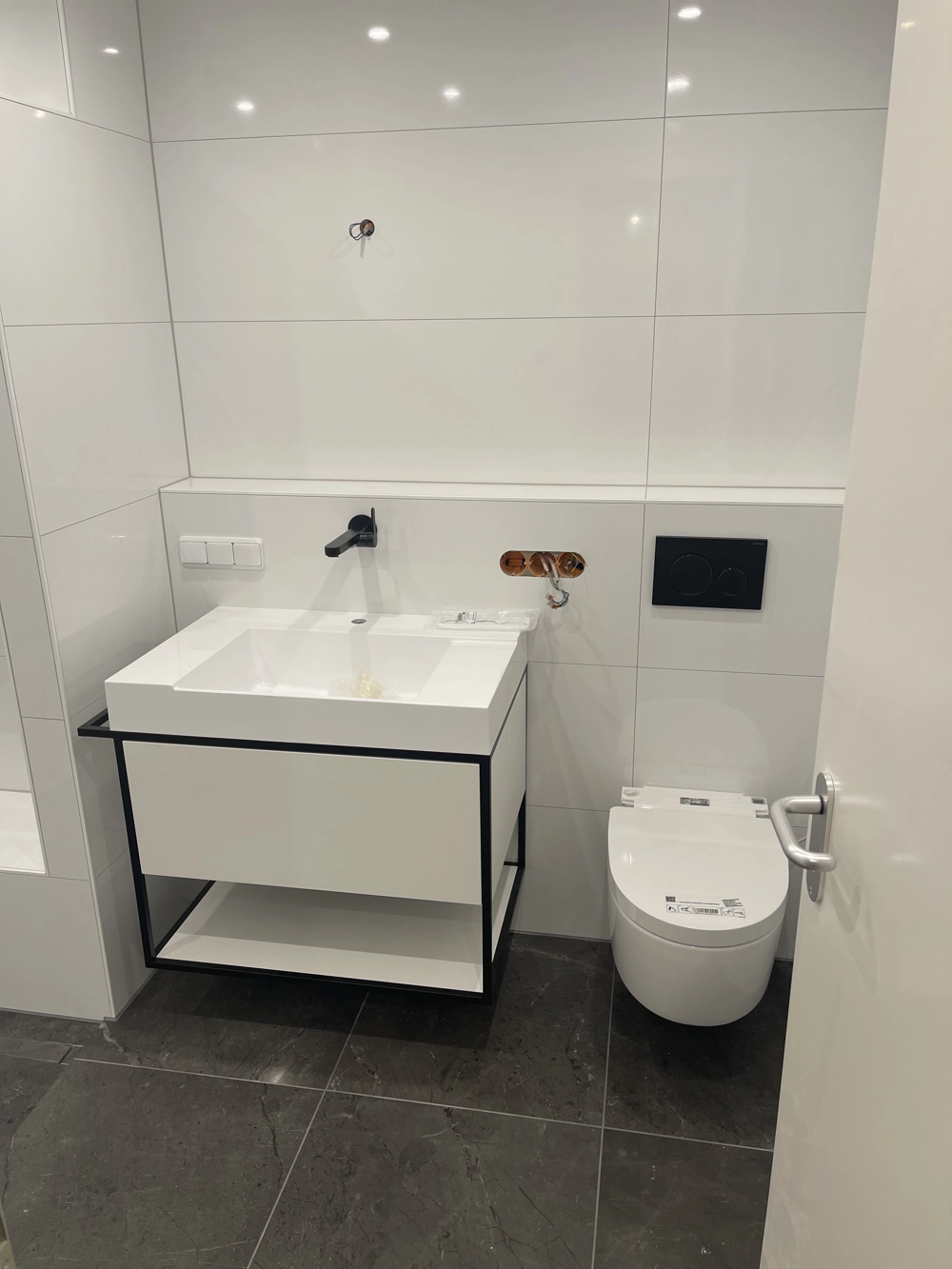 Modernes Badezimmer mit weißen Wand- und dunklen Bodenfliesen, schwarzem Waschtischgestell und wandhängendem WC nach Badrenovierung