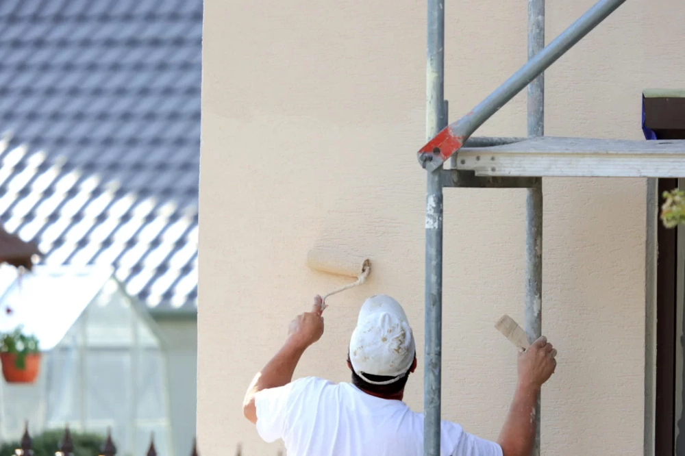 Maler trägt helle Fassadenfarbe mit Roller auf Außenwand auf neben Gerüst