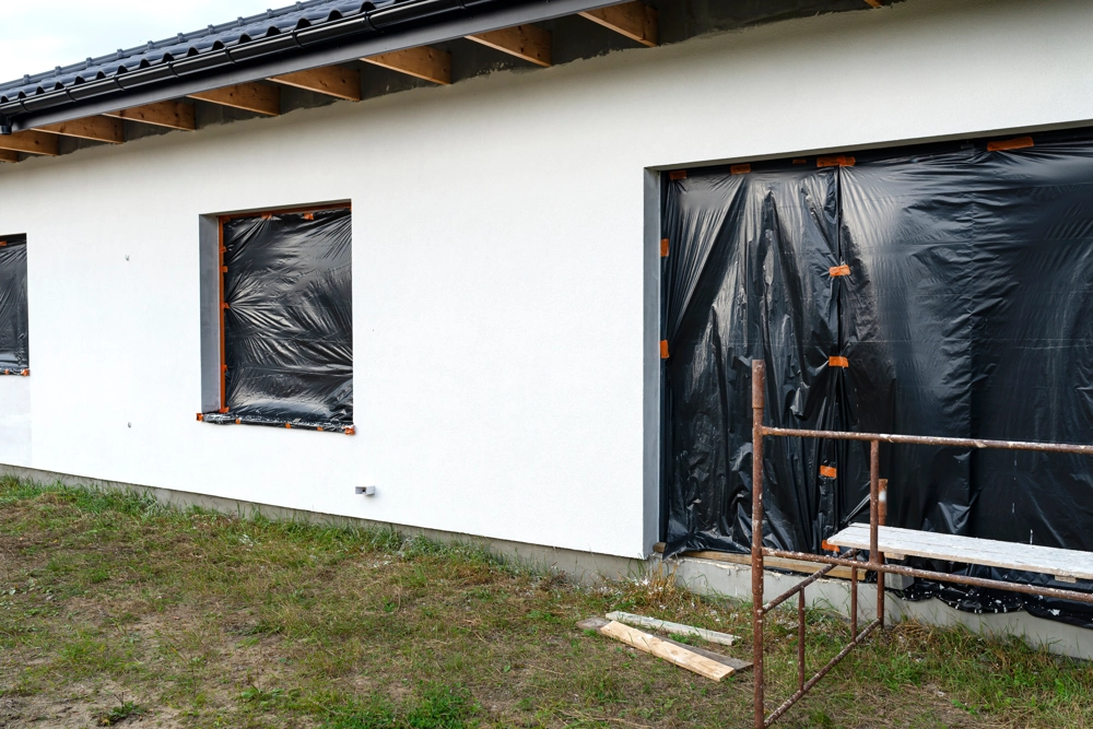 Weiß verputzte Hausfassade im Rohbau mit abgedeckten Fensteröffnungen und Gerüst – Putzarbeiten