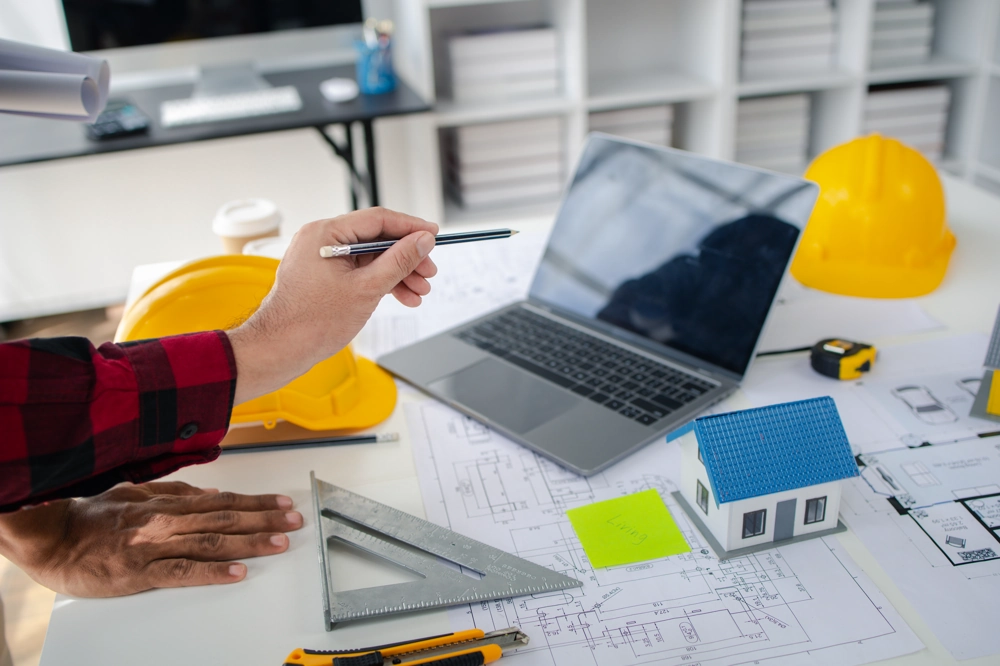 Heller Arbeitstisch mit Bauplänen, Laptop, Modellhaus, Lineal und Hand symbolisieren professionelle Bauplanung