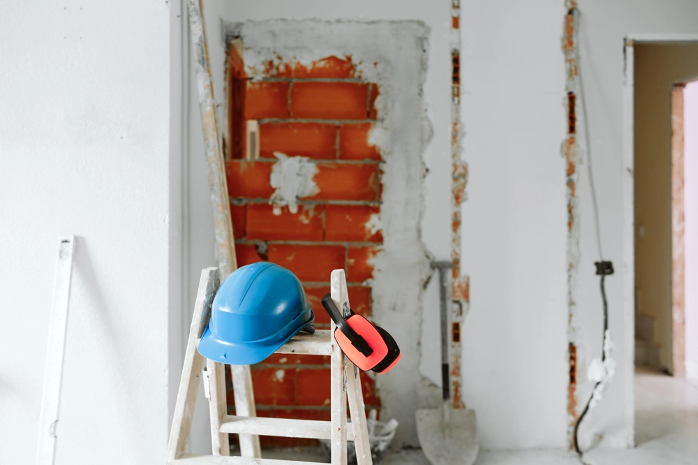 Innenansicht einer Baustelle mit Leiter, Schutzhelm und Gehörschutz vor freigelegter Ziegelwand