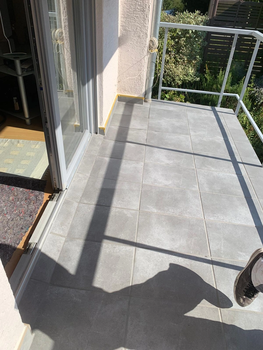 Balkon mit neu verlegten hellgrauen Fliesen auf Boden, weißem Geländer und Glasschiebetür – Fliesenleger