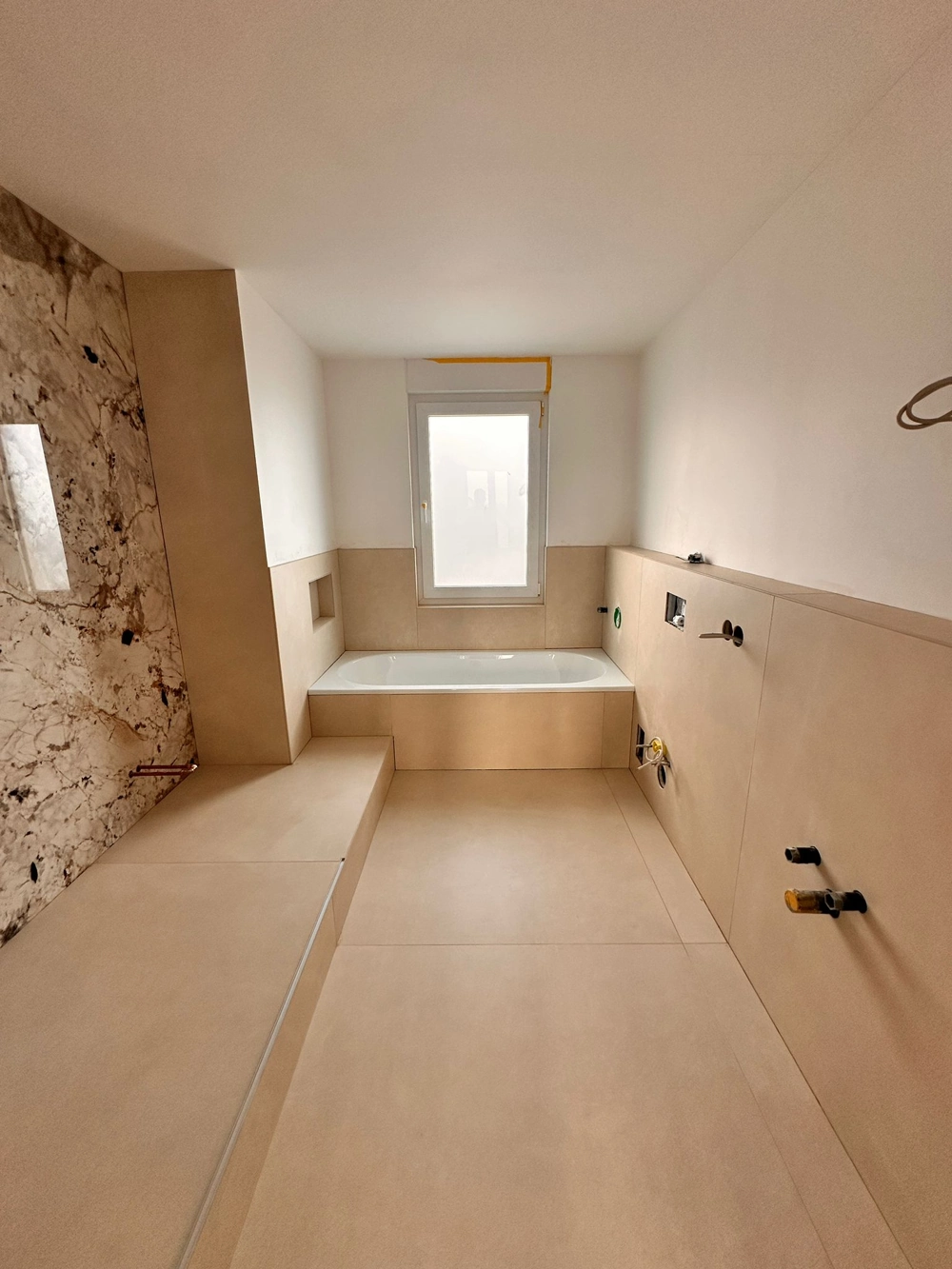 Badewanne mit beige Großformatfliesen an Boden und Wand vor weißem Fenster während Badrenovierung