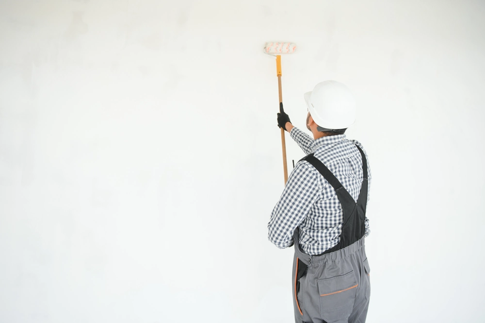 Handwerker mit Helm und Farbrolle streicht weiße Innenwand im Rahmen der Malerarbeiten