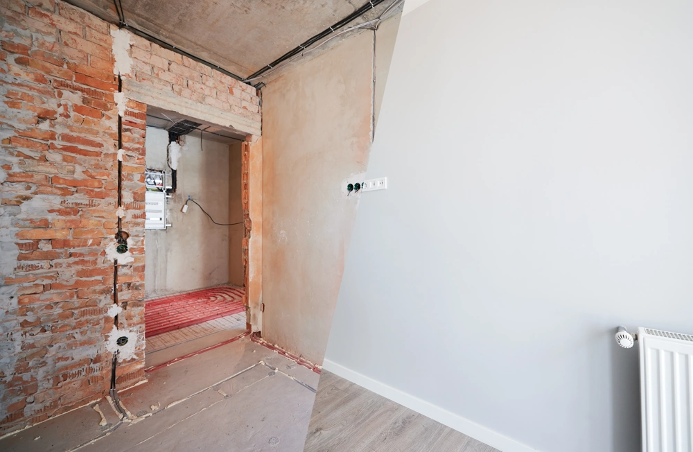 Halb entkernter Raum mit freigelegter Ziegelwand, Unterputz, freiliegenden Heizungsrohren und neuem Bodenbelag – Wohnungsrenovierung