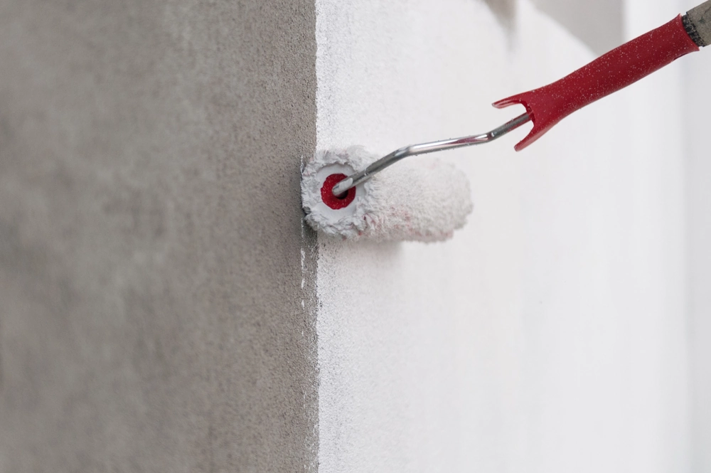 Farbrolle trägt weiße Farbe auf Wand auf, zeigt den Übergang von ungemalter grauer Fläche zu frisch gestrichener Wand – Malerarbeiten