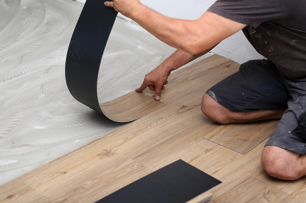 Handwerker kniet auf Boden und verlegt Vinylboden-Planke mit schwarzem Rücken auf Klebstoff, Vinylboden verlegen
